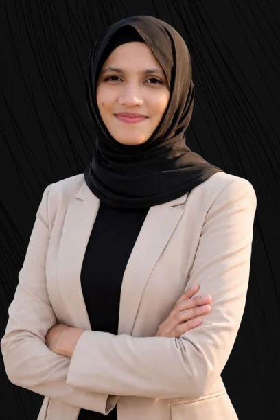 A young woman wearing a black hijab and beige blazer stands with folded arms, smiling softly at the camera against a black background. Digital Marketing Freelancer in Ernakulam